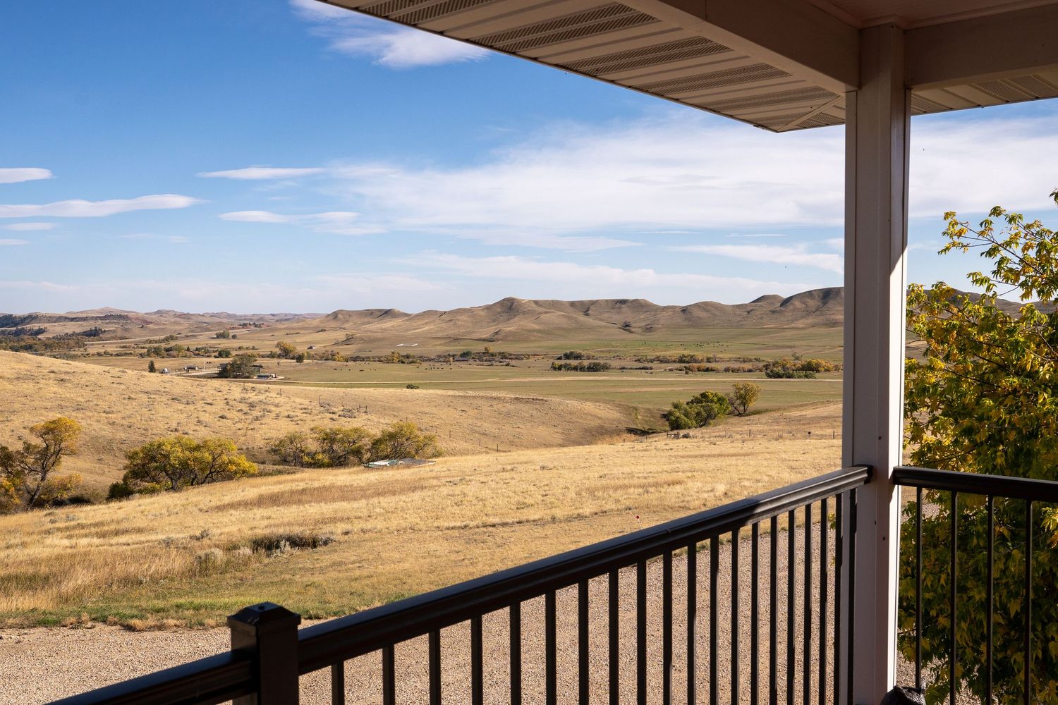 A porch with wooden railings overlooks vast golden grasslands and rolling hills under a bright blue sky.