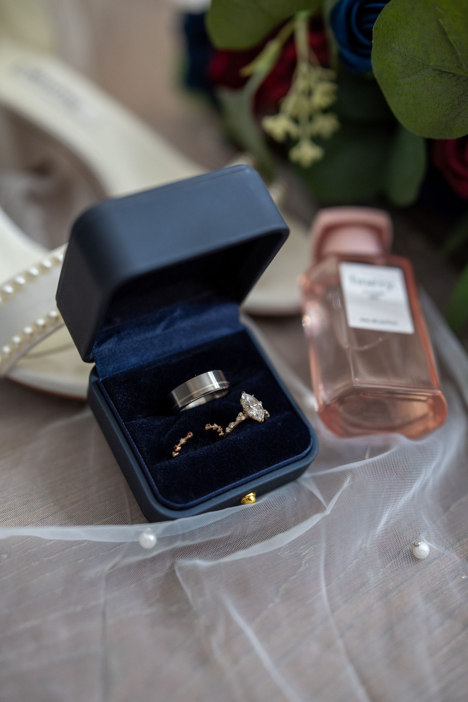 Wedding rings in navy blue box with perfume bottle and floral decor.