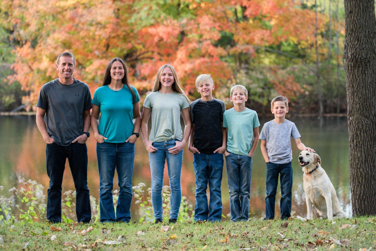 Family poses in line against autumn trees for outdoor group portrait.