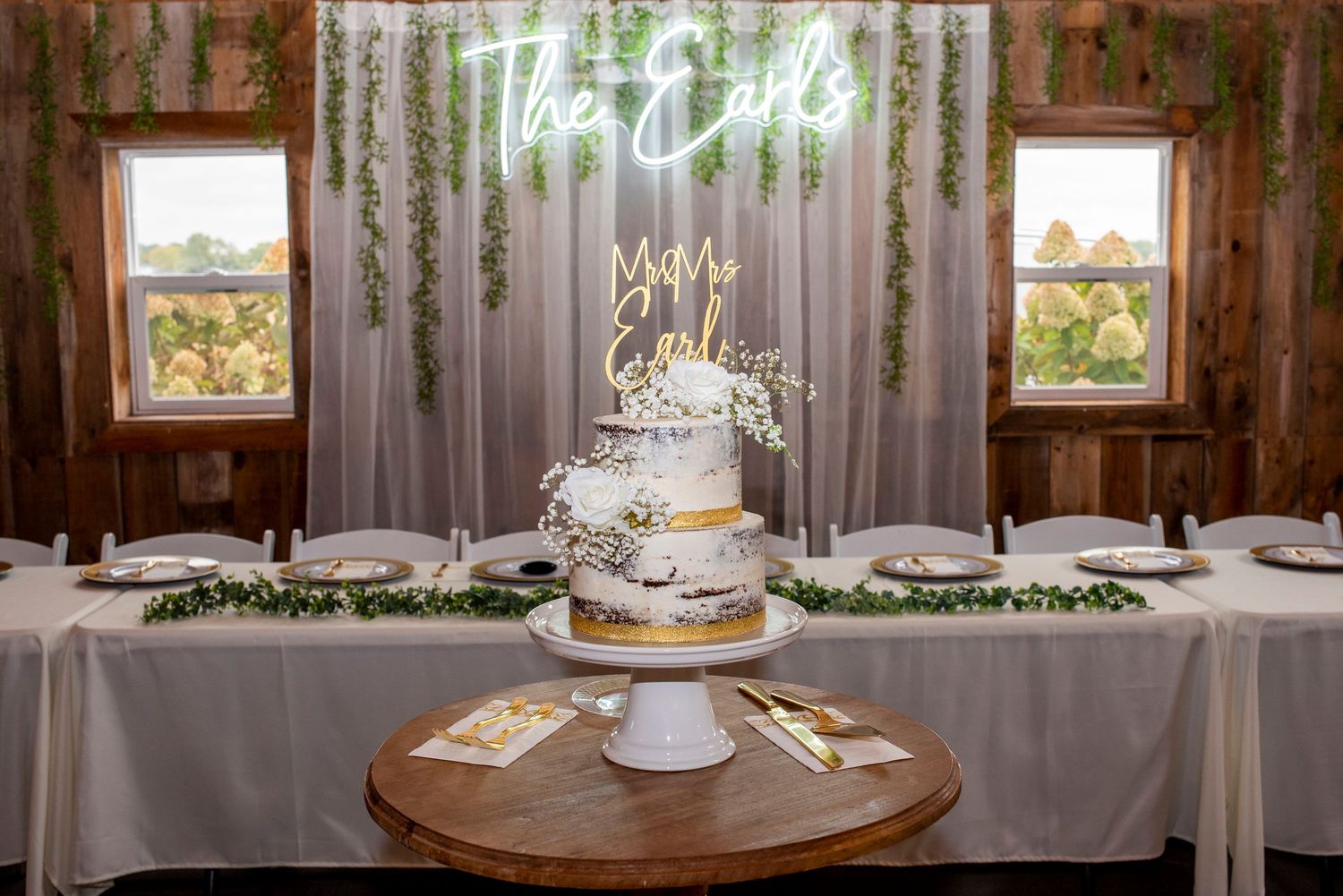 Elegant three-tier white wedding cake displayed on wooden table with neon sign and windows in rustic venue.