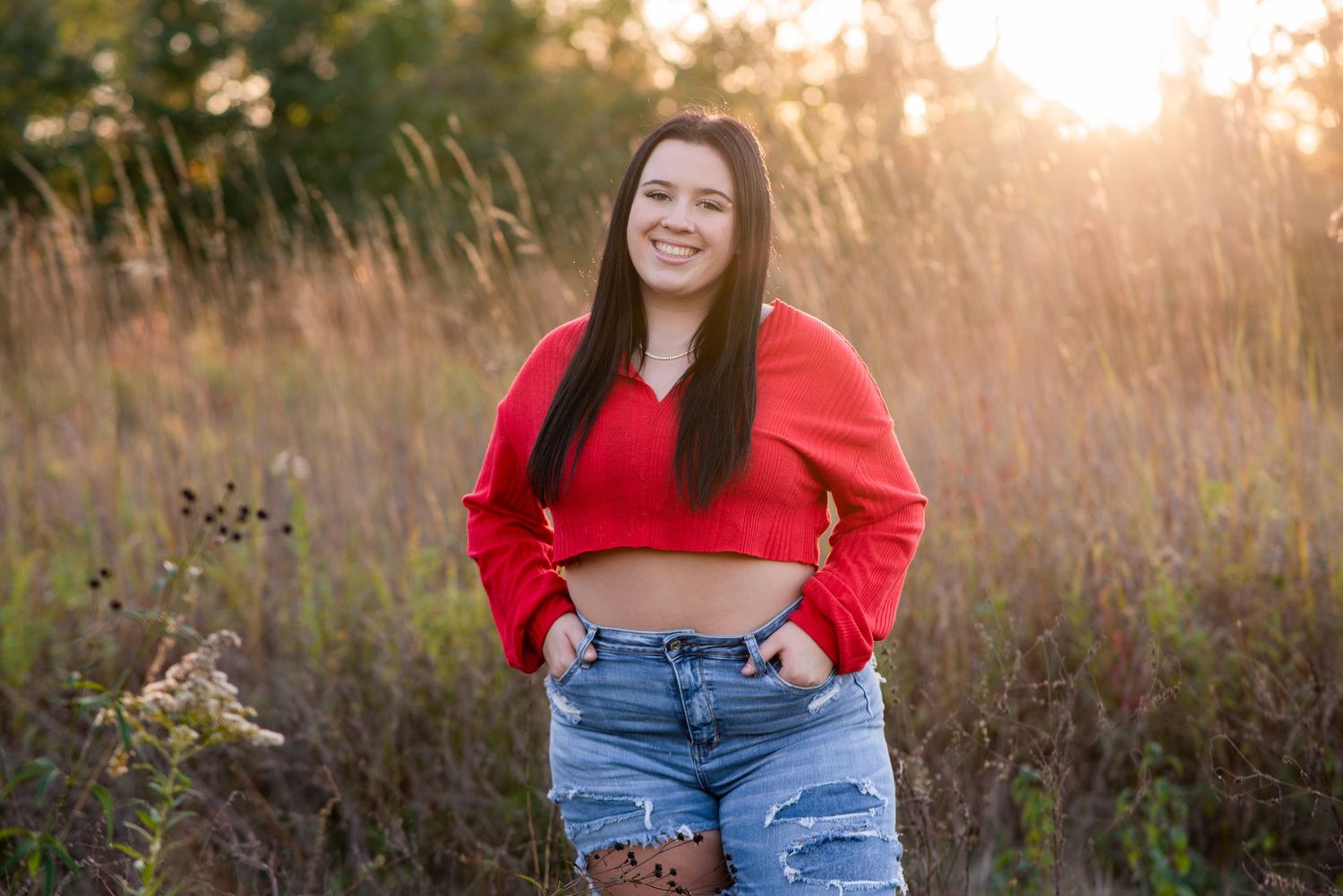 Young woman in red crop top and denim shorts posing in golden field at sunset.