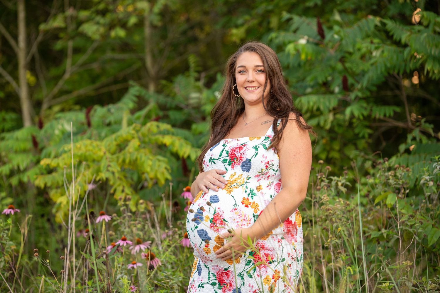 A woman in a floral dress poses outdoors in a garden setting with lush greenery and flowers in the background.