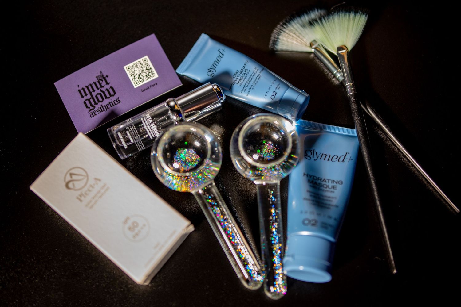 A collection of beauty and cosmetic products including makeup, skincare items, and brushes arranged on a dark surface.