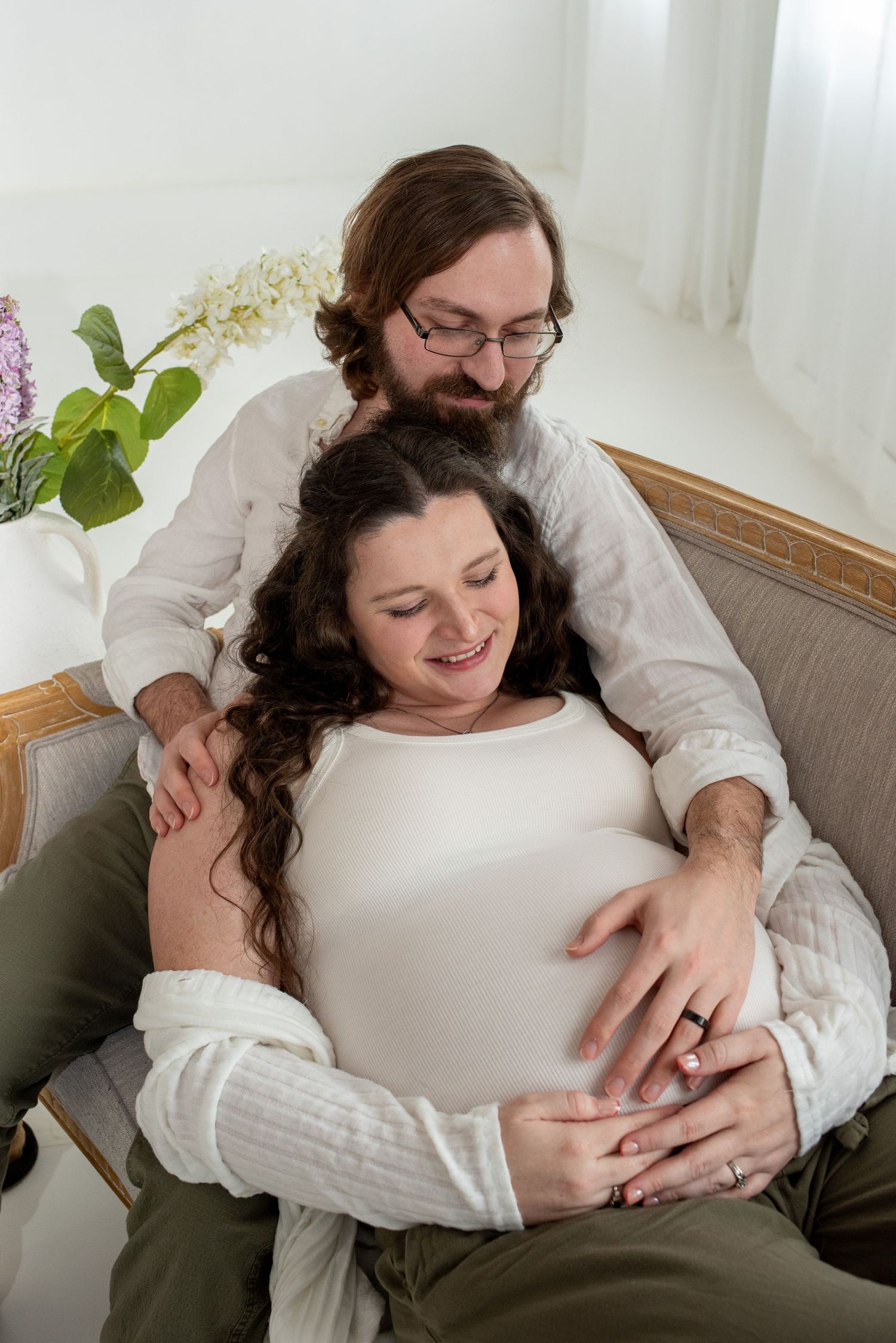 A couple in white clothing embraces while sitting on a couch during an intimate maternity photoshoot.