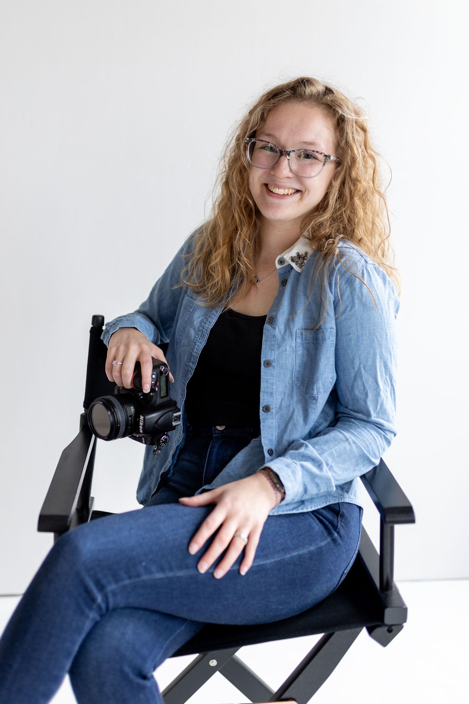 Portrait session showing photographer with camera sitting casually in director's chair against white backdrop.