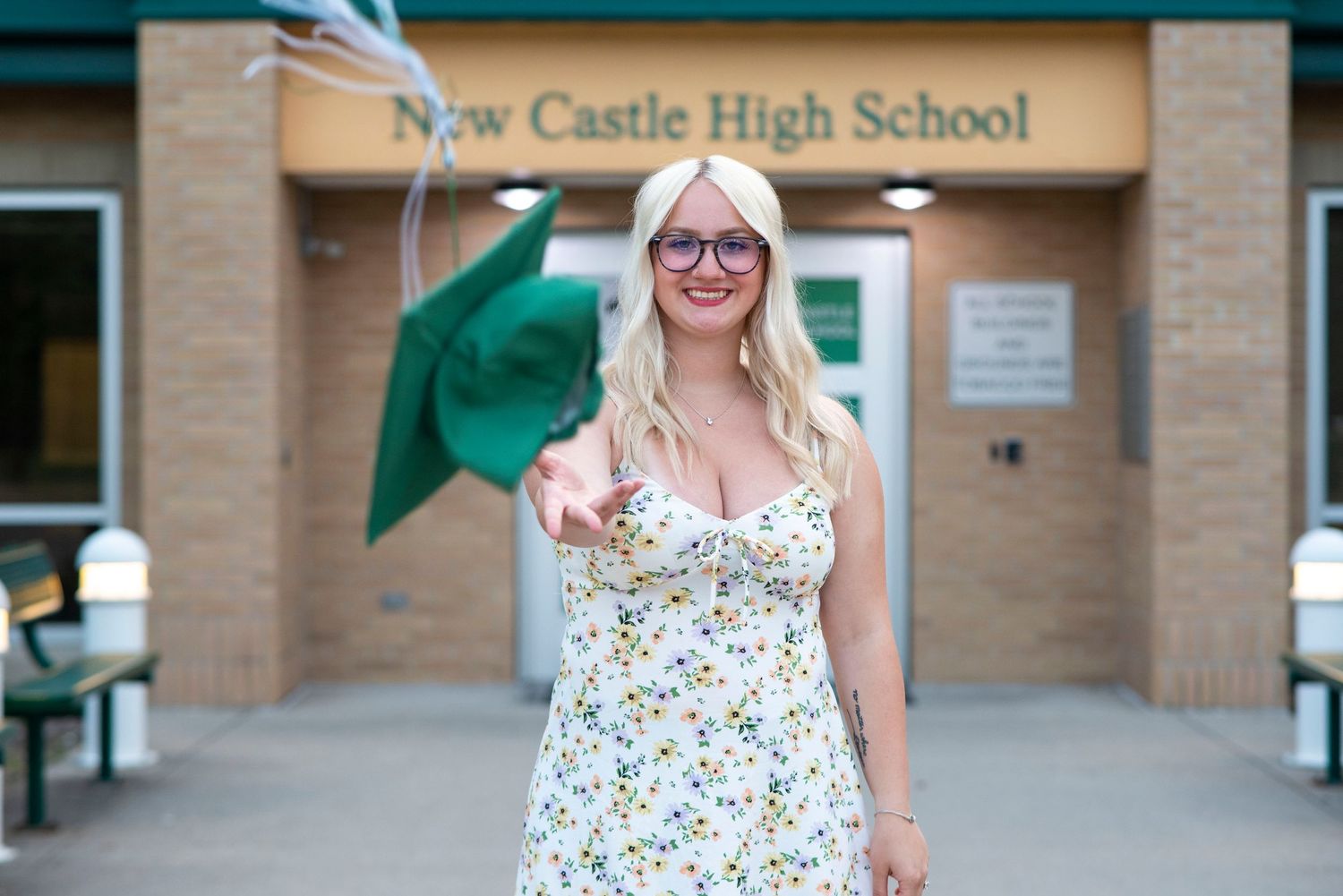 Graduate in floral dress and glasses stands outside New Castle High School entrance with graduation cap.