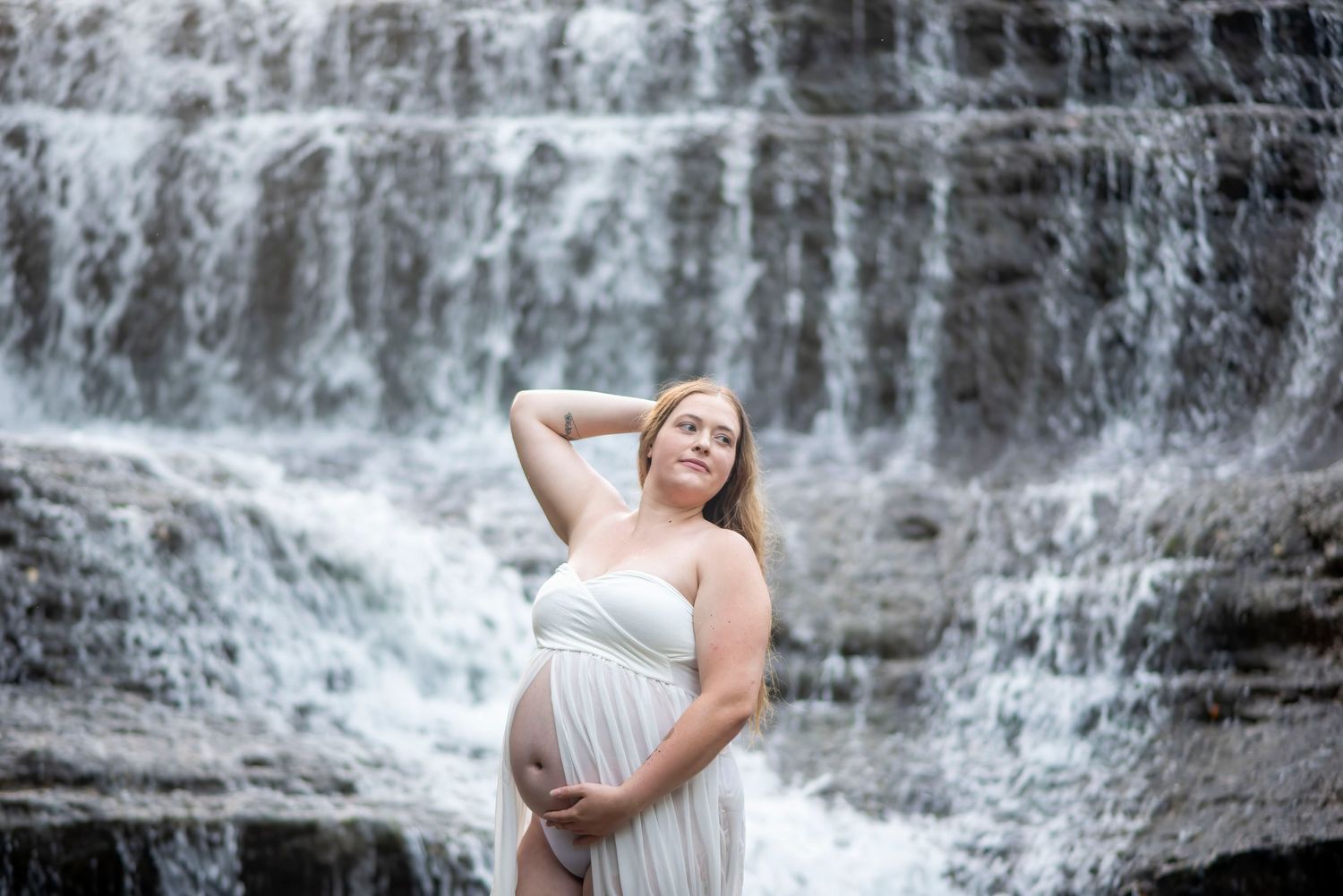 Maternity portrait of an expectant mother in white dress standing before a cascading waterfall.