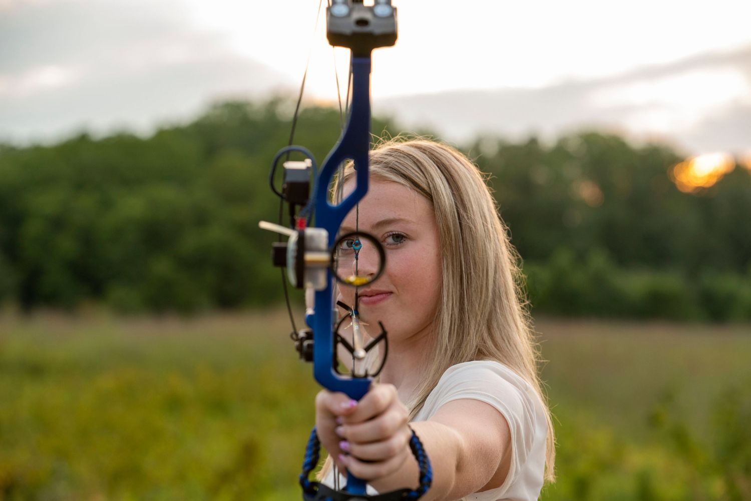 Person practicing archery with a compound bow in an outdoor field at sunset.