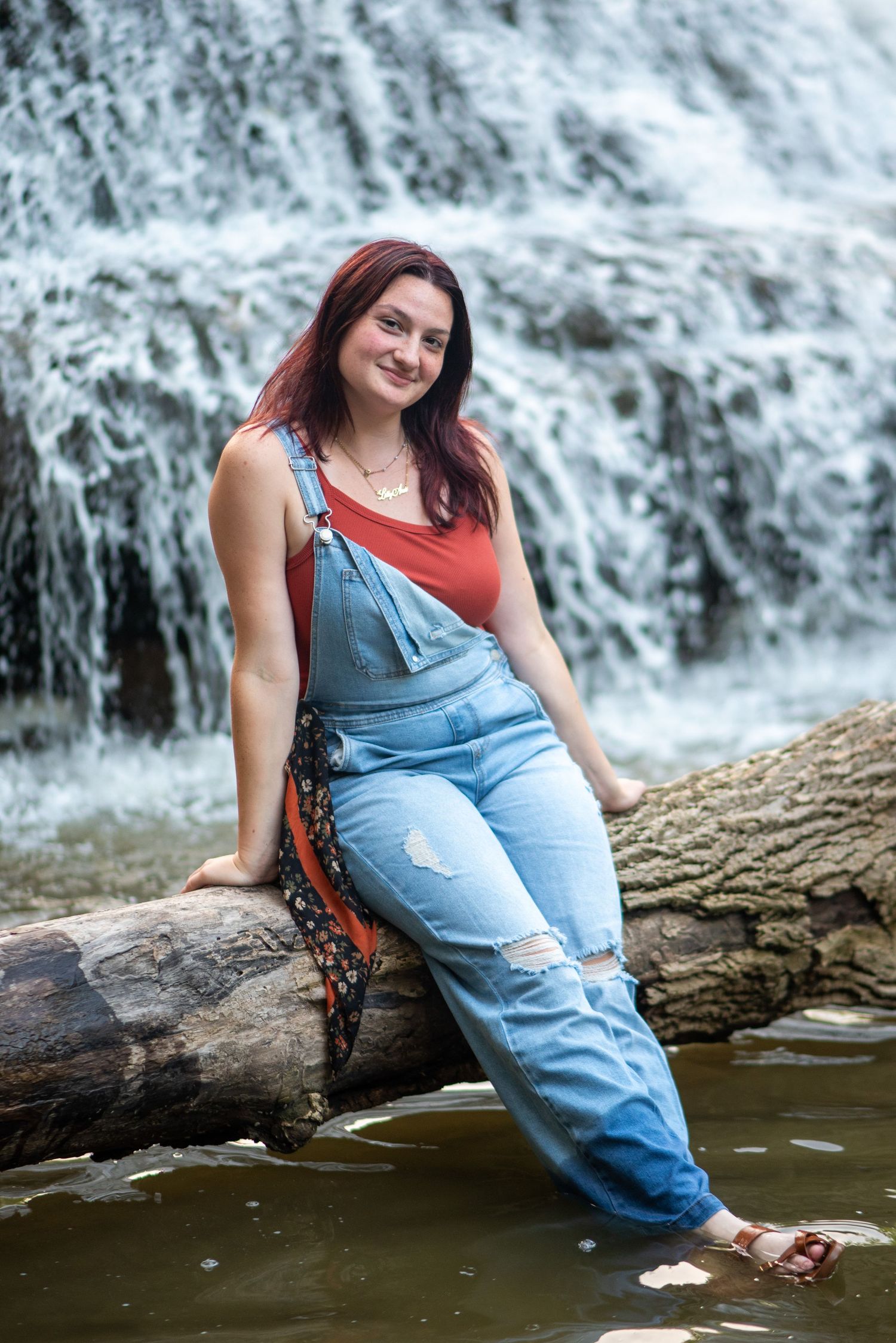 Portrait session by a waterfall featuring someone in distressed denim overalls and red top.