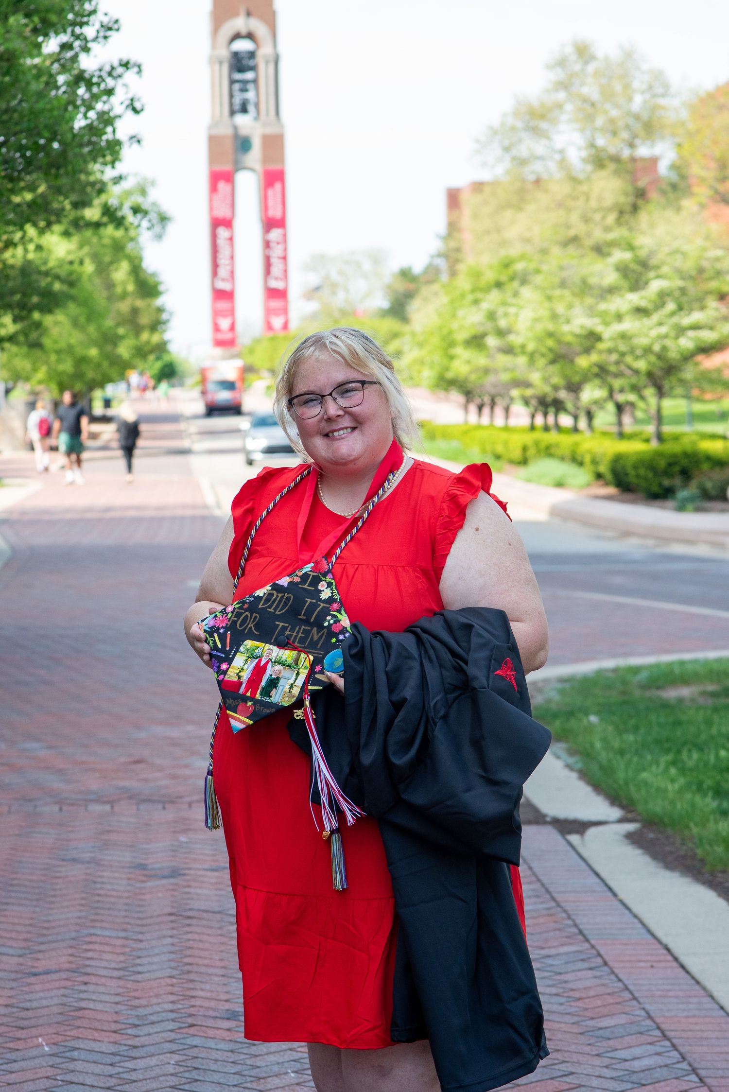Graduate in red and black academic regalia displaying their graduation tassel on a sunny campus walkway.