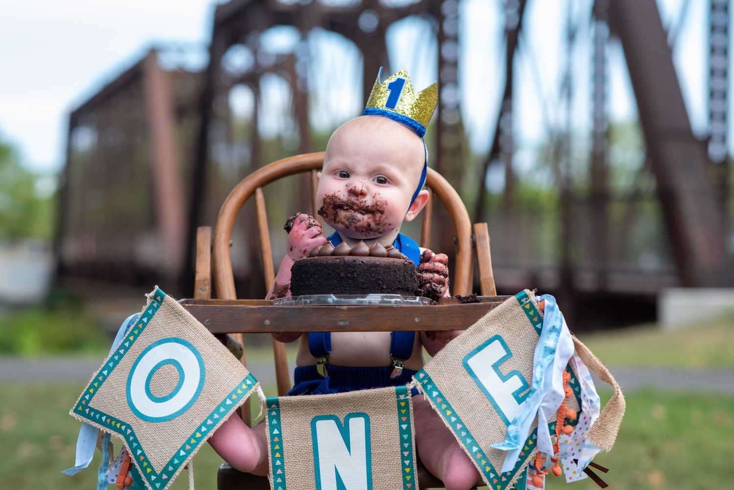 Baby in high chair with blue 'ONE' banner celebrating first birthday with messy cake.