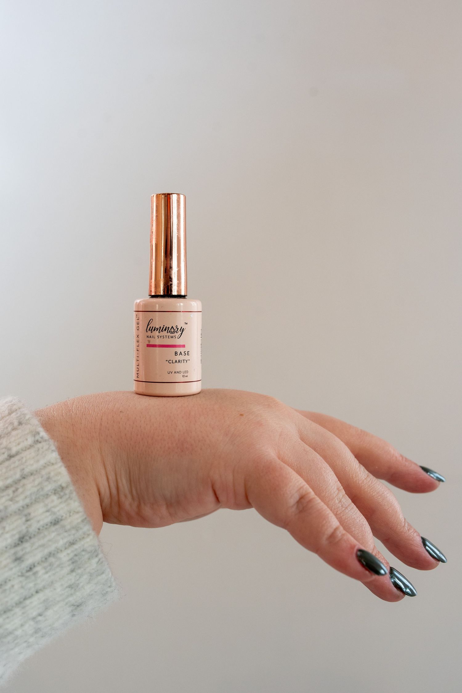 A hand with black nail polish holds a rose gold beauty product bottle against a light marble background.