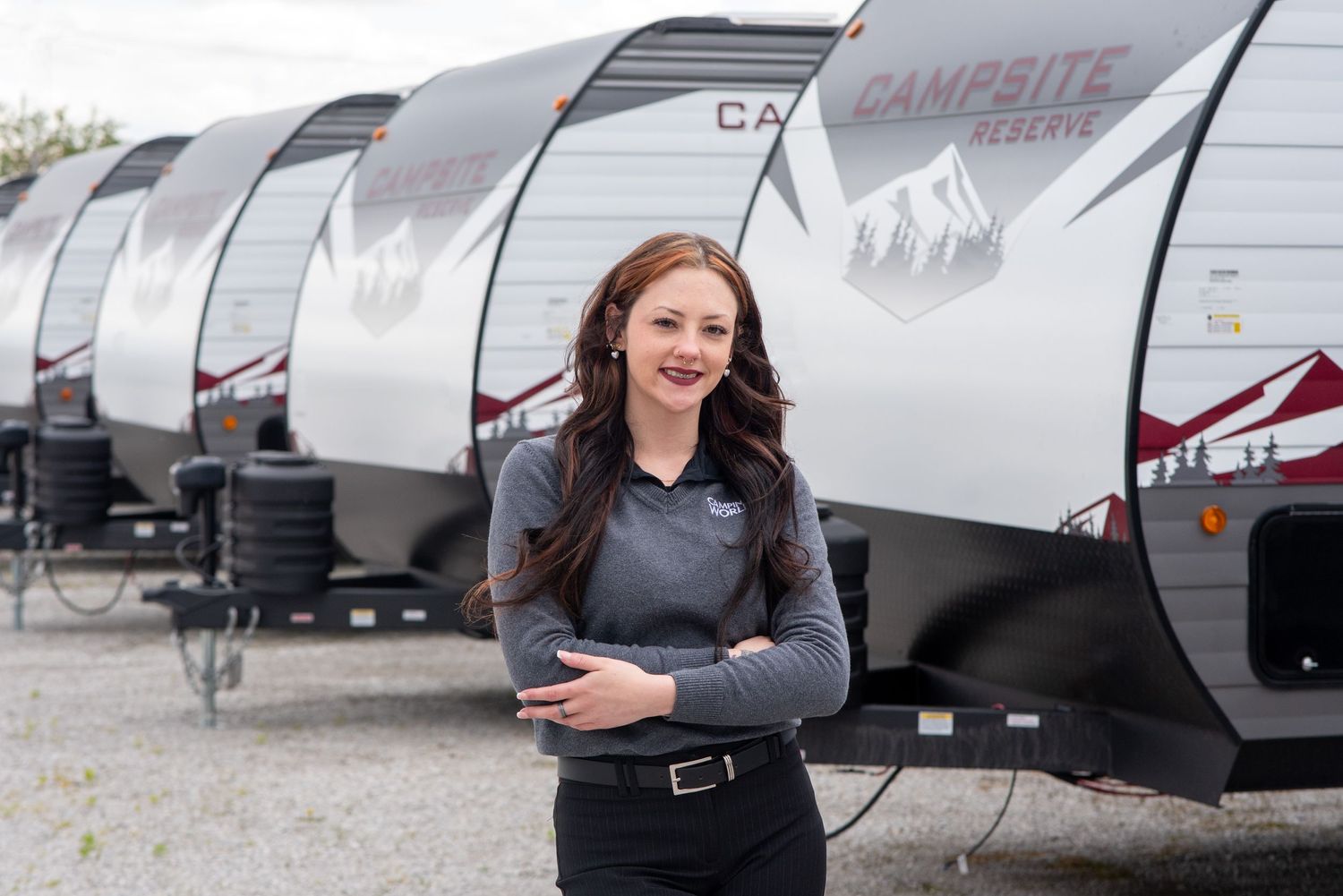 Woman in casual attire poses beside white travel trailers in outdoor setting.
