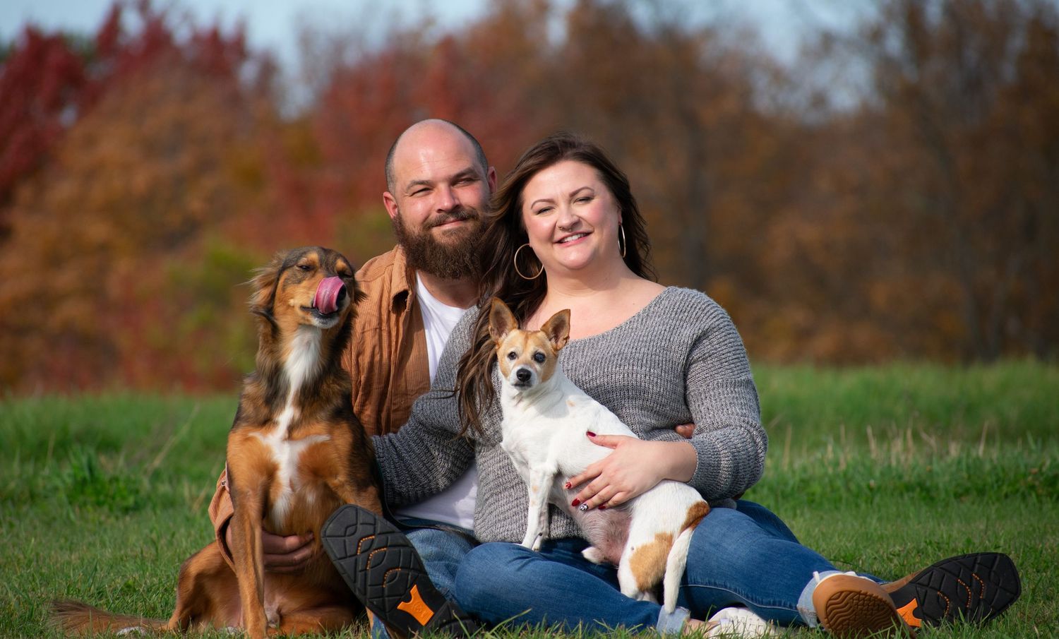 A couple and their dogs pose together in a scenic fall setting with colorful foliage.