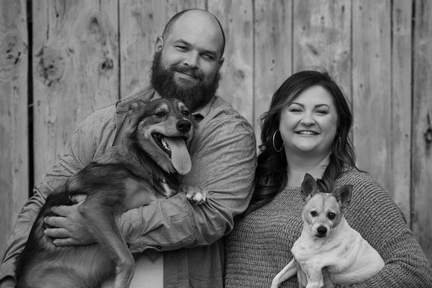 Black and white portrait of couple with their two rescue dogs against wooden wall.