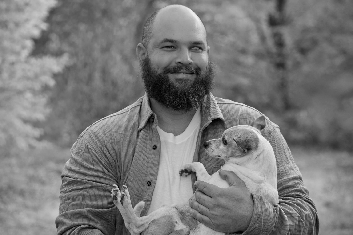 Man in casual attire holds small dog while smiling outdoors.