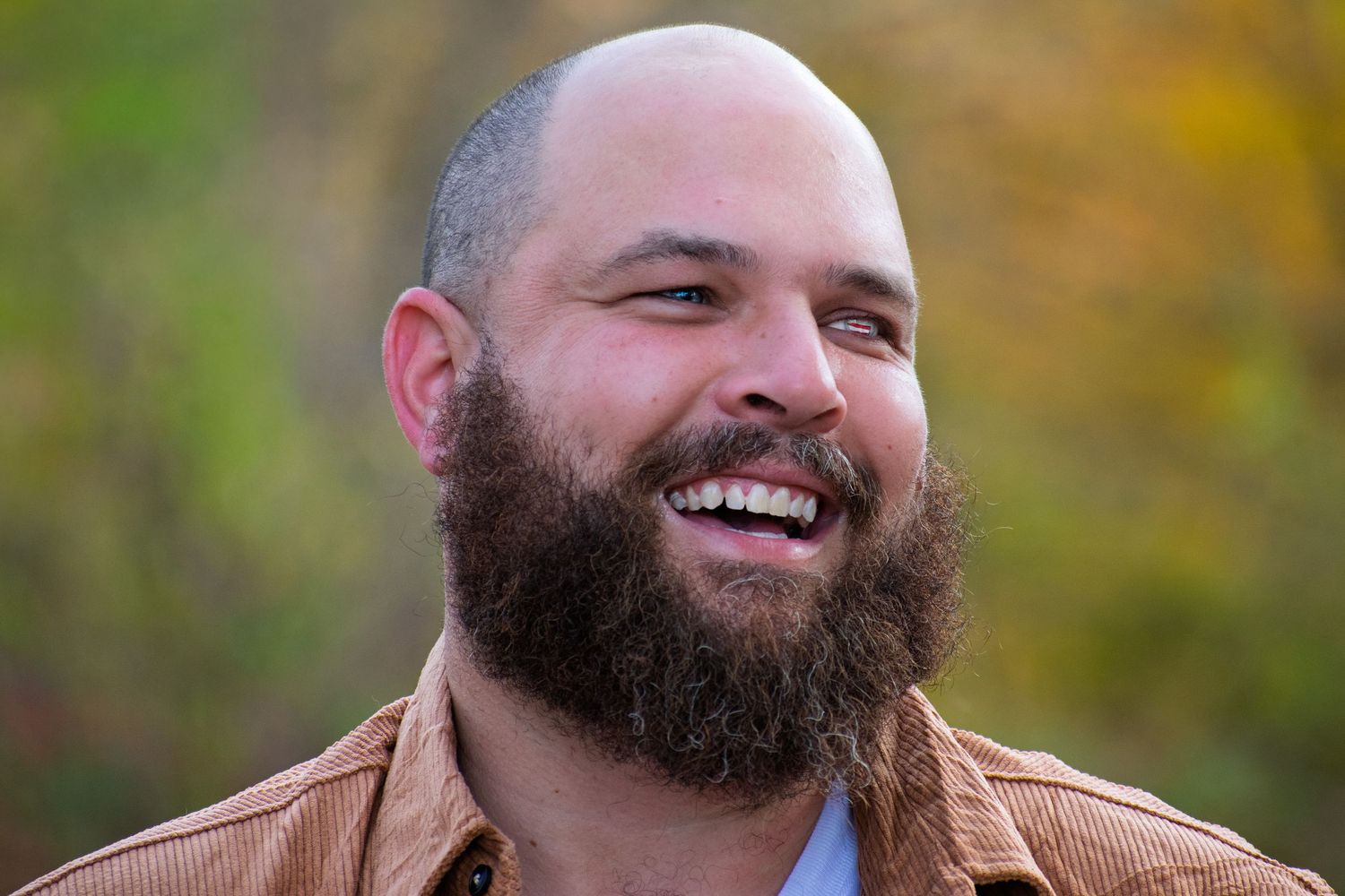 Man with full beard smiles warmly in outdoor autumn setting.