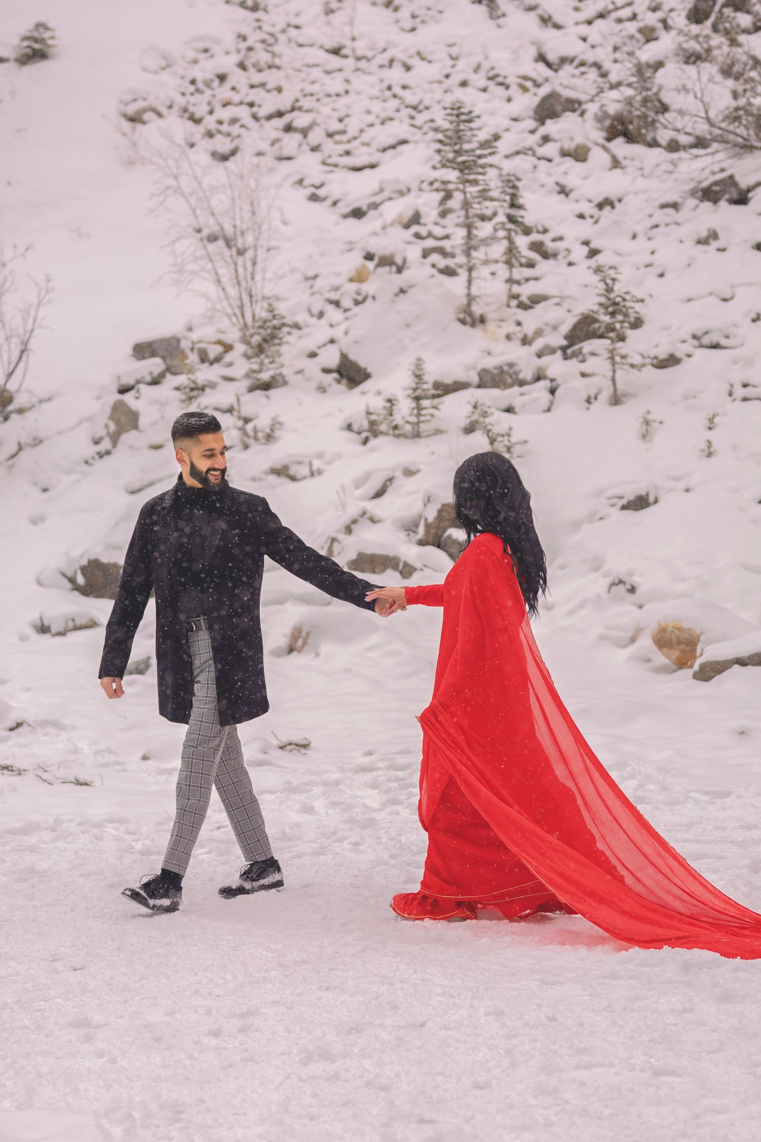 Couple holding hands in snowy landscape, woman in flowing red gown, man in black coat, romantic winter photoshoot.