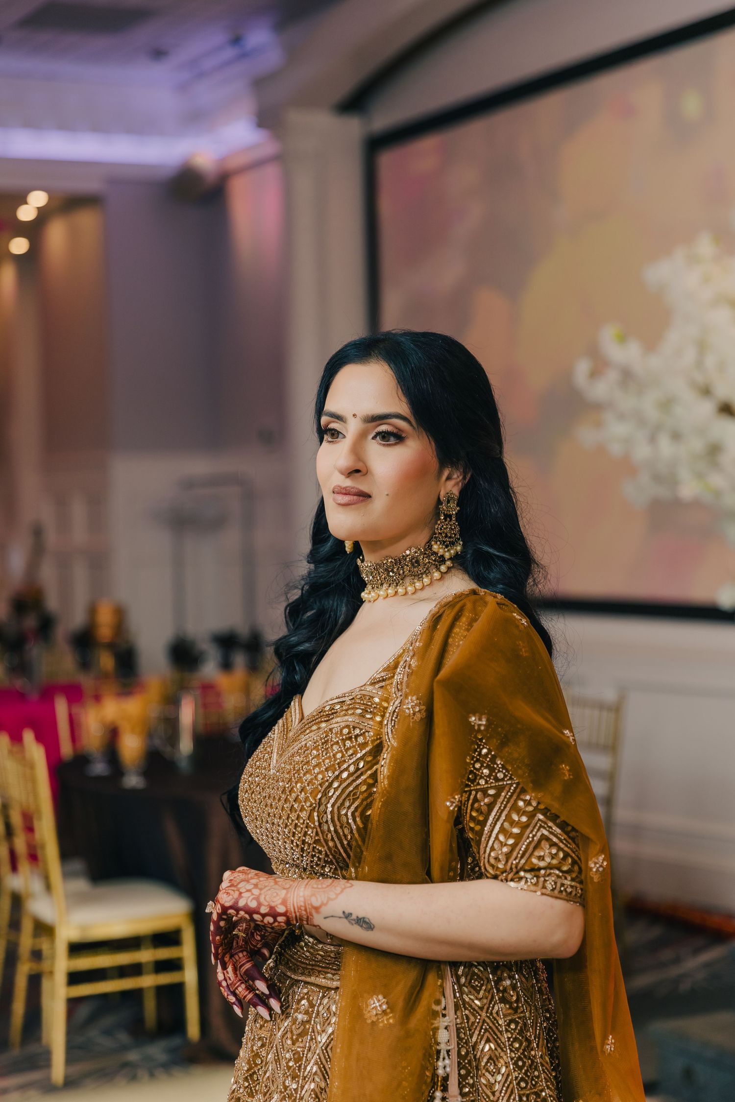 A woman in a mustard-colored traditional Indian lehenga with intricate embroidery and gold jewelry stands at a wedding reception.