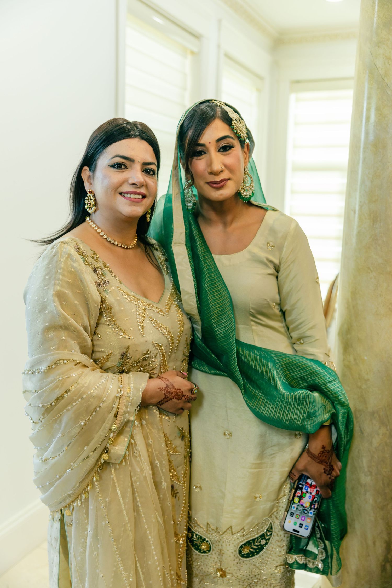 Two women in elegant traditional South Asian attire pose together in a bright indoor setting.
