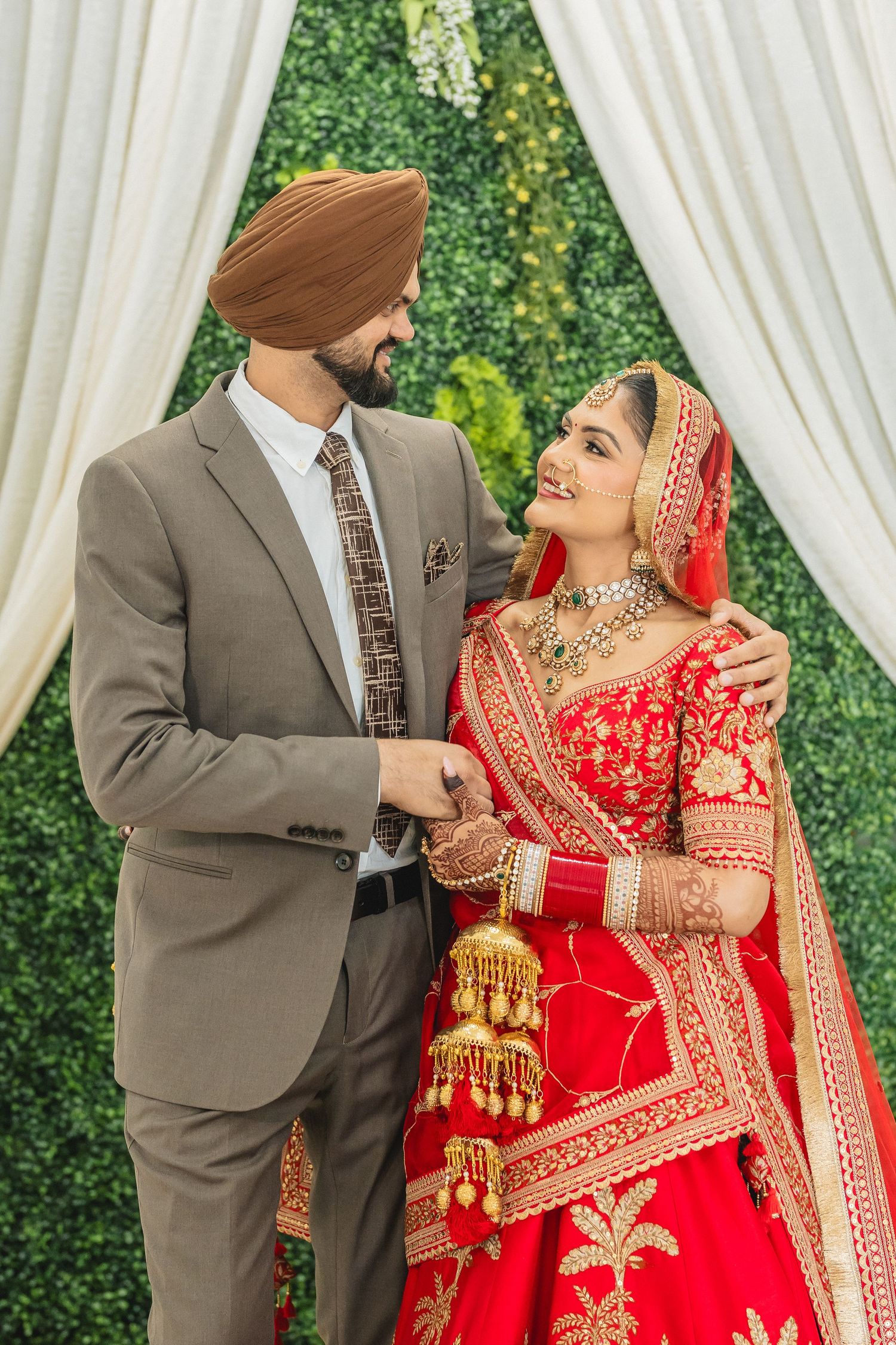 A couple in traditional Indian wedding attire share romantic moments in a garden setting with white curtains.