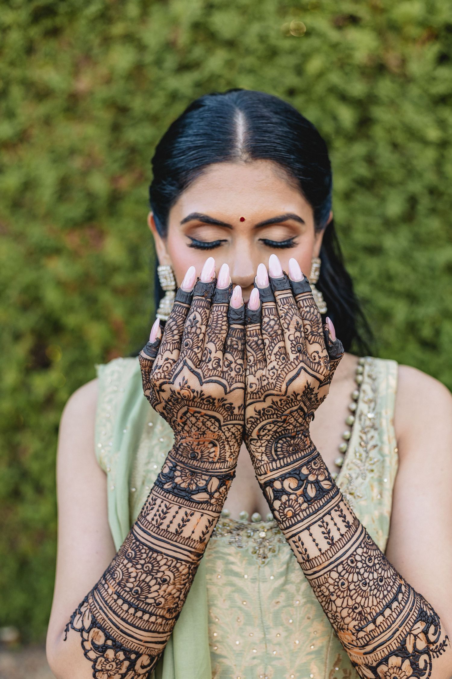 Intricate henna mehndi designs displayed on hands against a light green traditional outfit background.