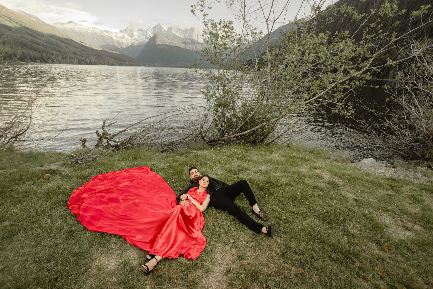 A couple poses on grassy shores with a dramatic mountain lake backdrop.