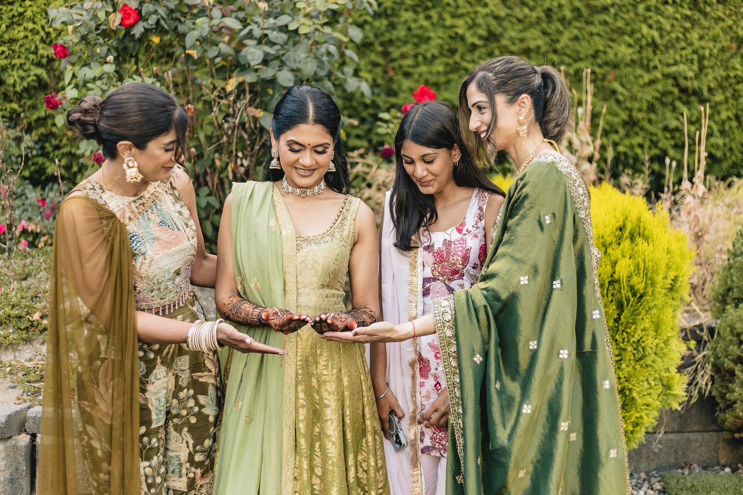 A group wearing traditional South Asian attire gathers outdoors in a garden setting with greenery and flowers.