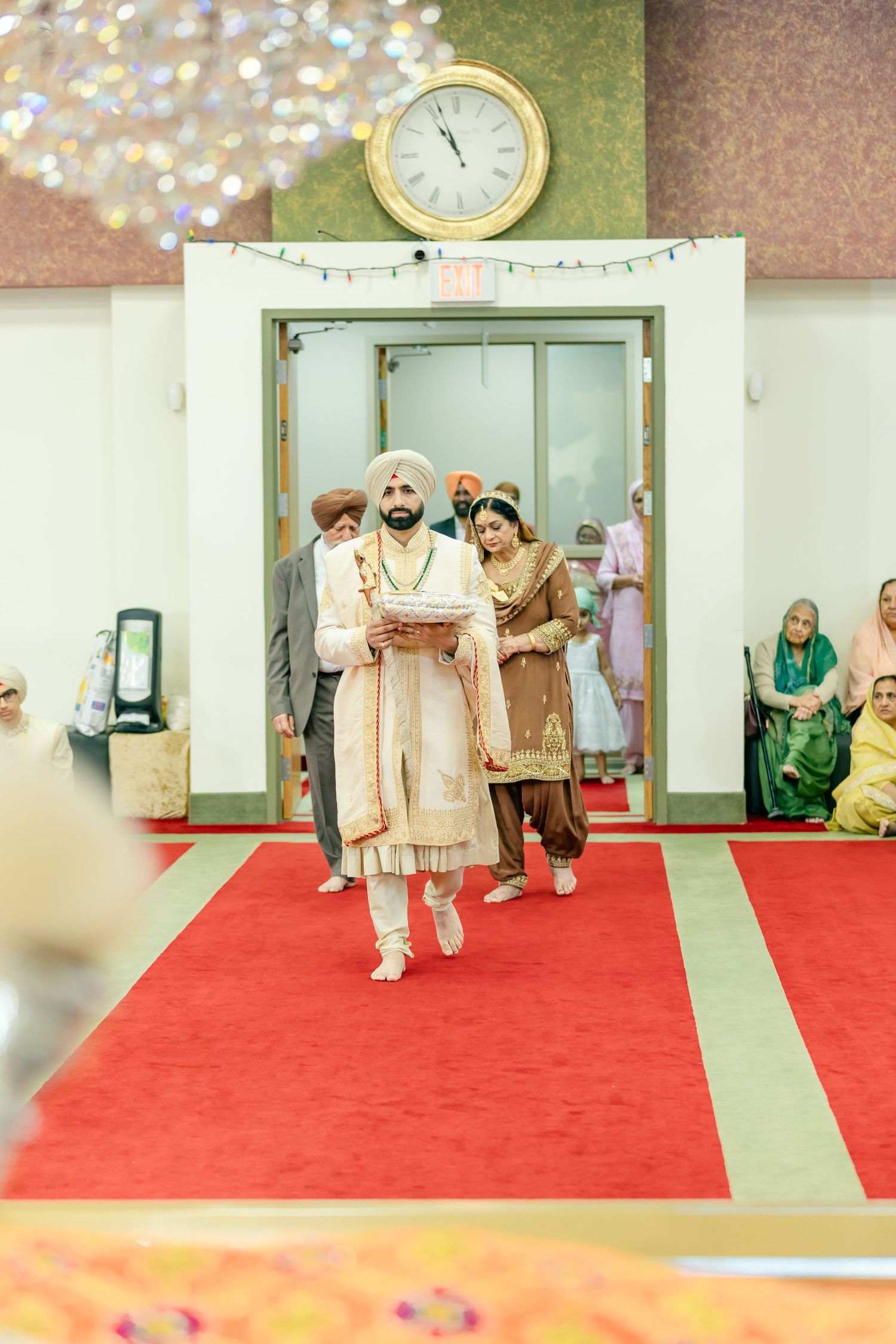 Wedding ceremony procession on red carpet in indoor venue with traditional decorations.