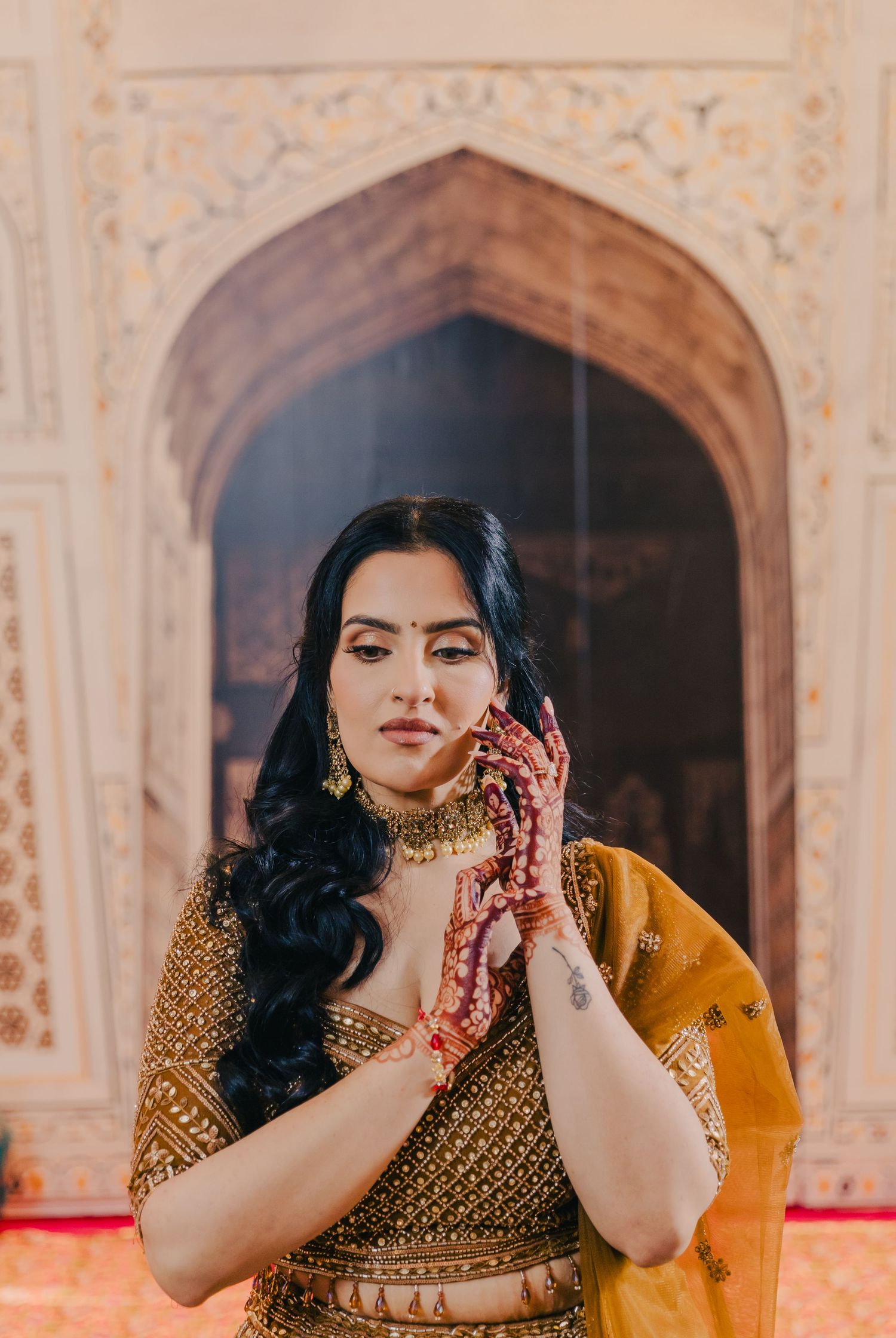 Woman in golden lehenga shows traditional henna designs on her hands against architectural backdrop.