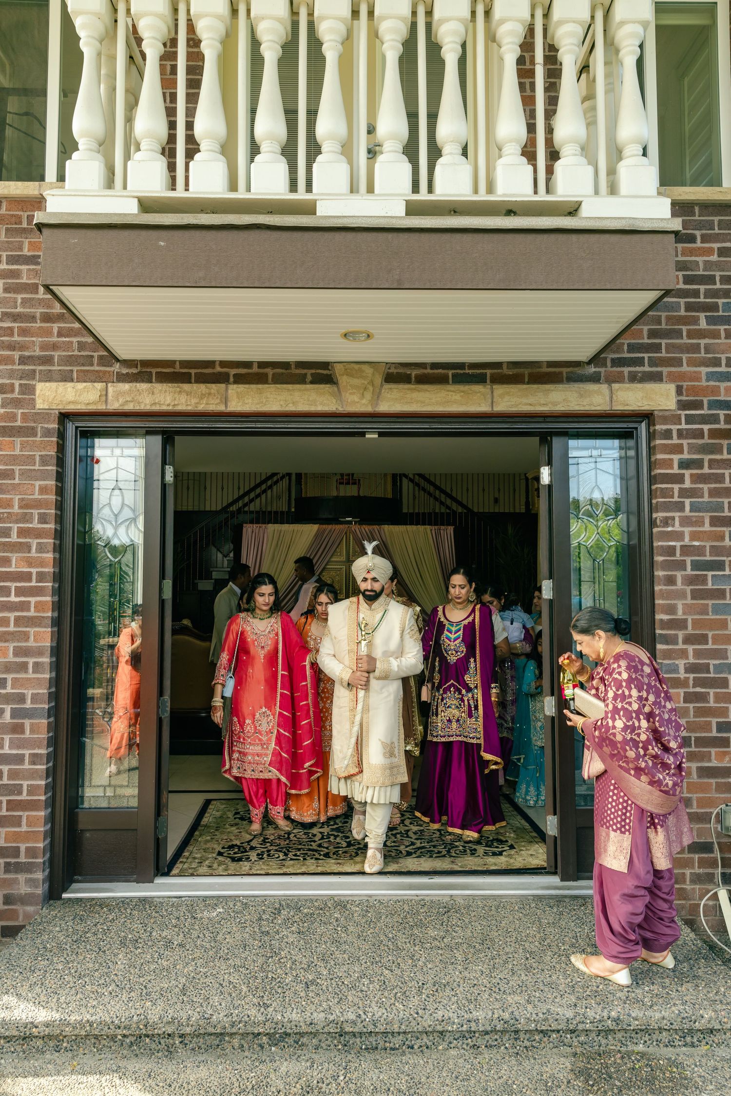 Wedding party in colorful traditional Indian attire gathered outside brick building entrance.