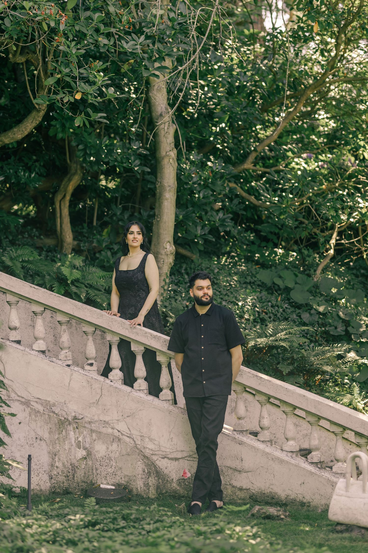 A couple poses along stone balustrades surrounded by lush green foliage.