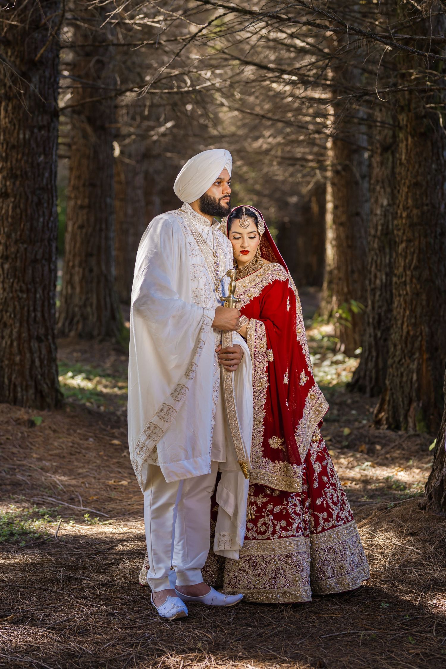 Couple in traditional Sikh wedding attire posing among pine trees in a forest setting.