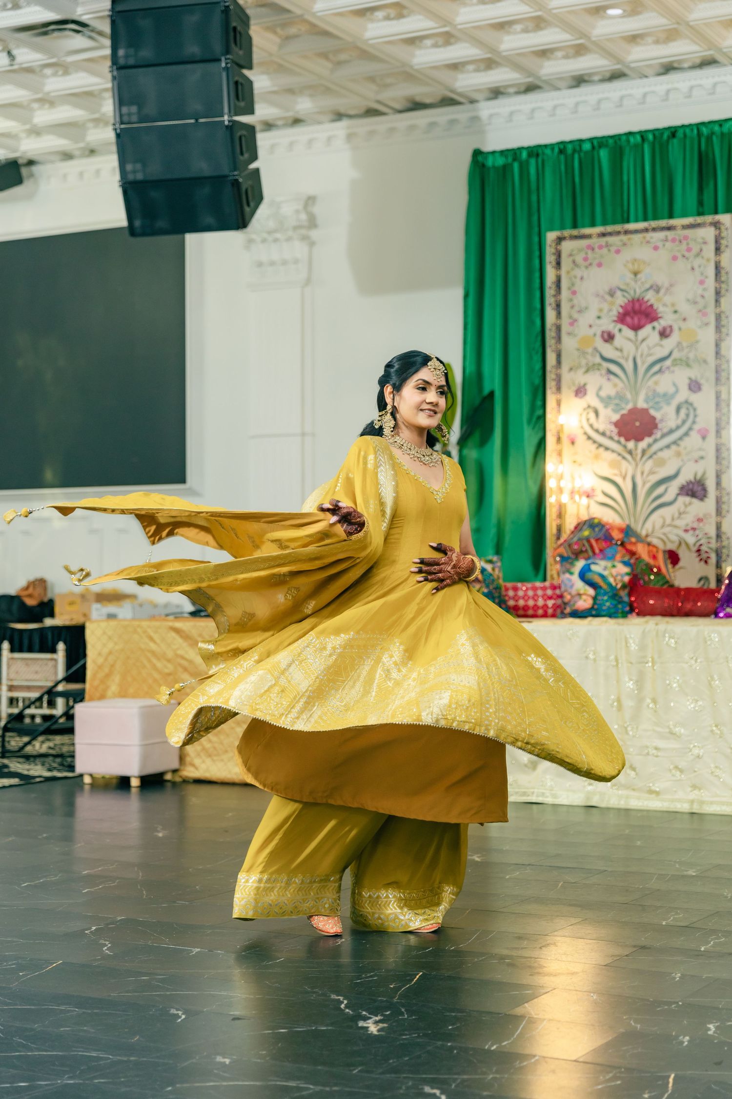 Dancer in flowing gold dress performs graceful spinning movements on dark floor with green backdrop.