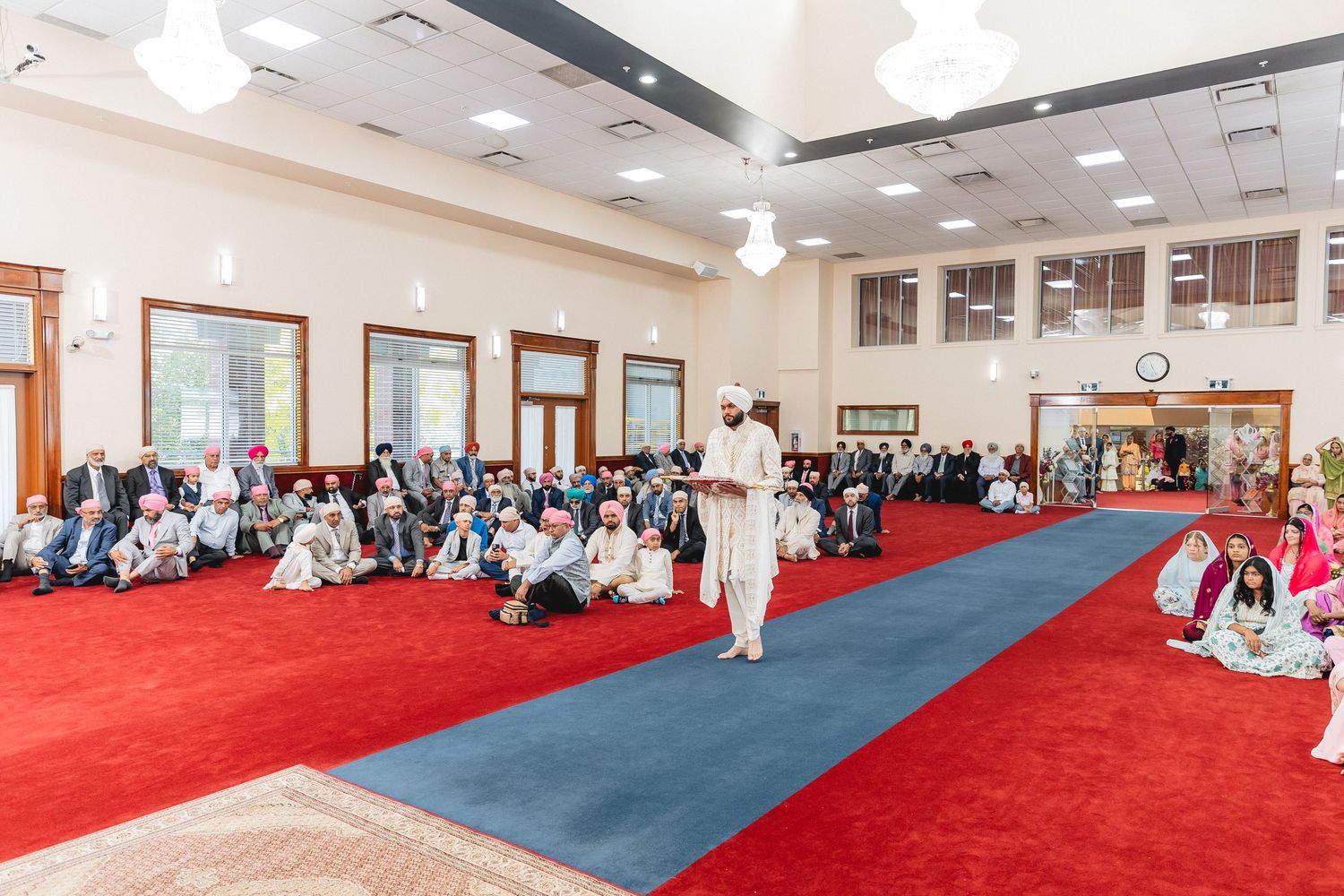 Students in white martial arts uniforms sit along red and blue mats during a karate class in a bright indoor studio.