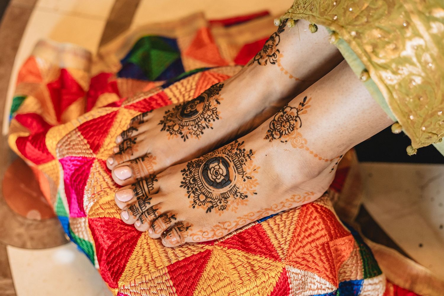 Intricate henna design adorns feet resting on colorful traditional textiles with geometric patterns and gold accents.