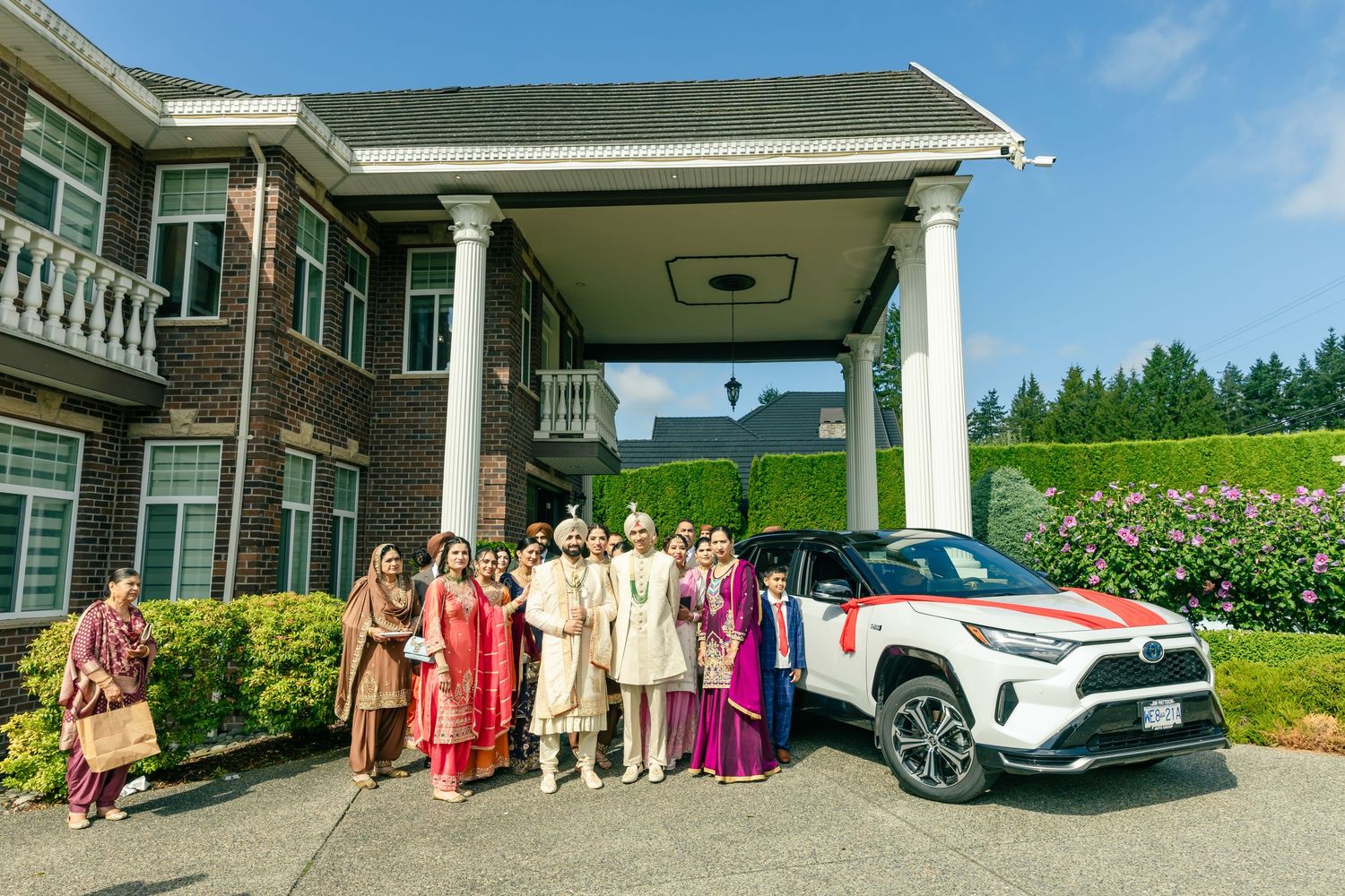 Wedding party members in formal attire gathered outside a large house with vehicles in background.