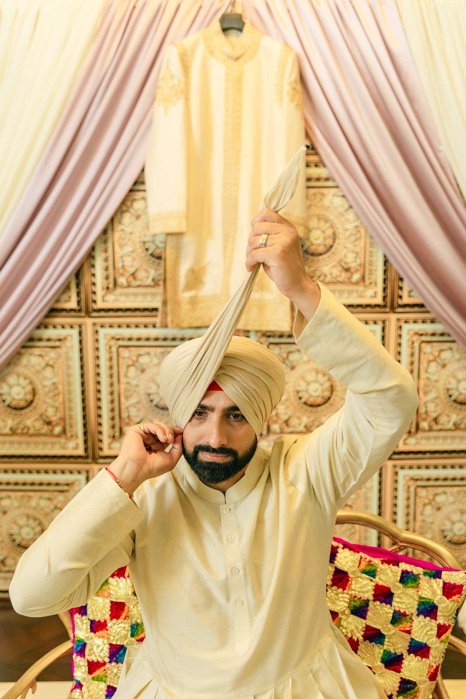 Person in cream colored traditional Sikh attire and turban poses playfully against ornate backdrop with drapes.