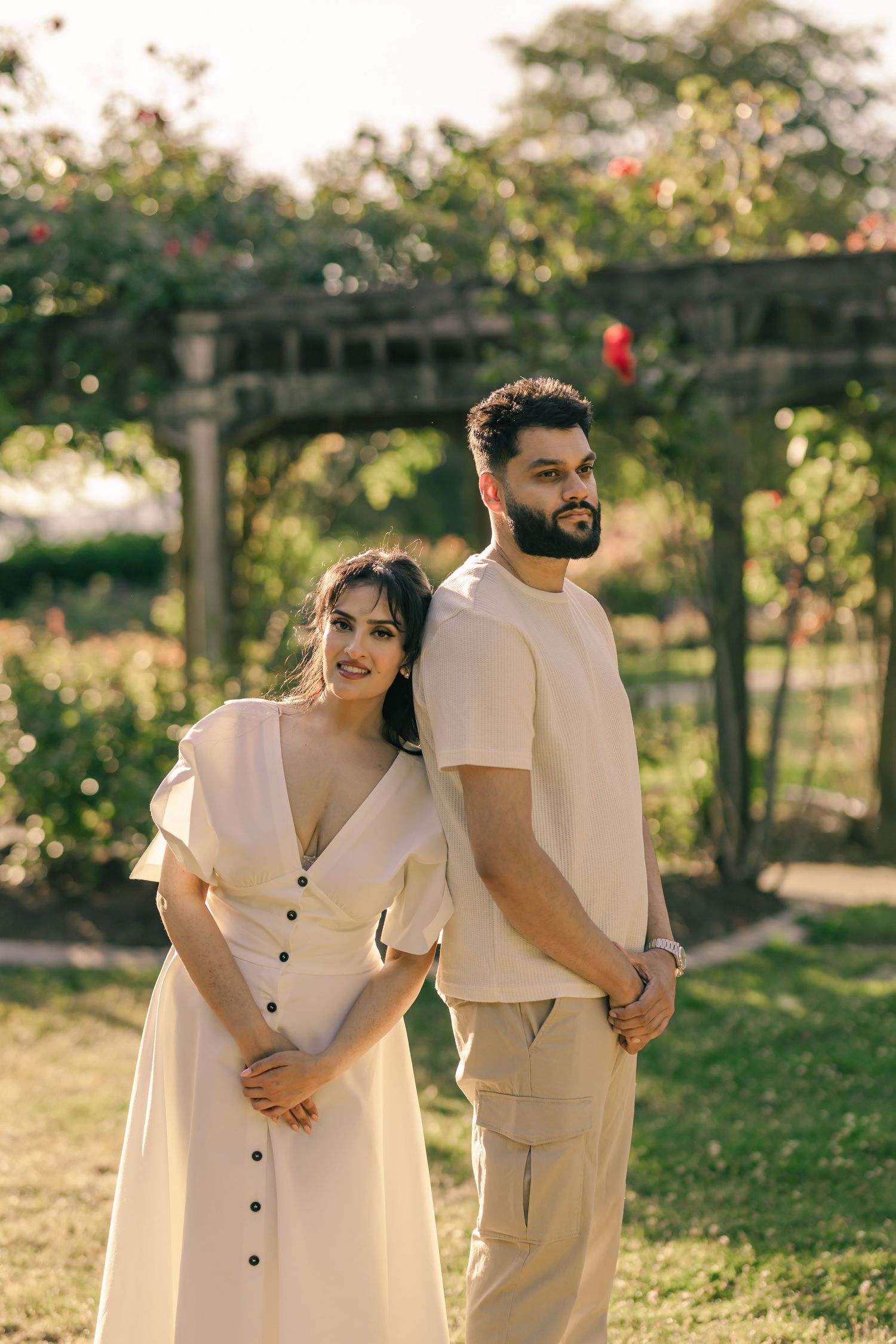 A couple in light-colored outfits poses in a garden setting during golden hour.