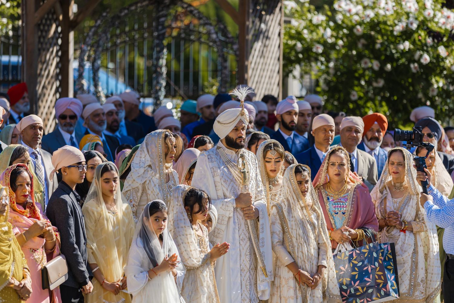 Large group wearing traditional white Indian wedding attire gathered outdoors in sunny garden setting.