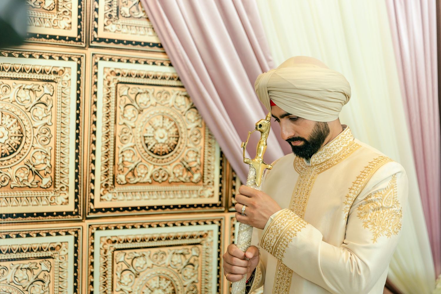Person in light colored traditional Indian attire adjusts their turban against decorative wall backdrop.