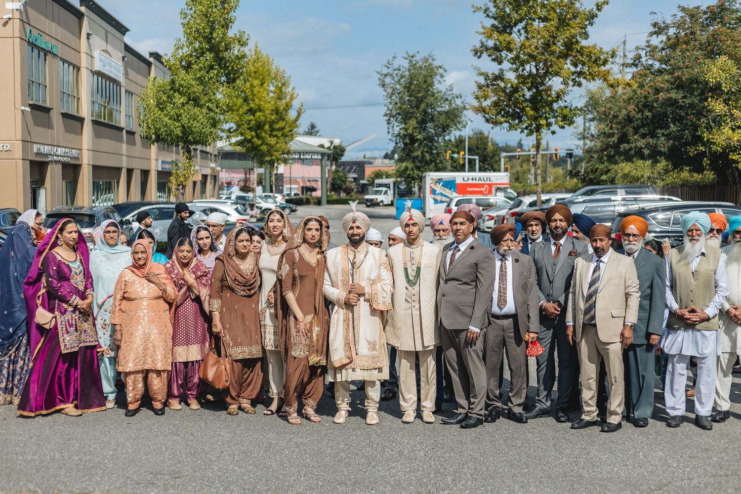 Large group of wedding guests in formal attire posed together outdoors on a sunny day.