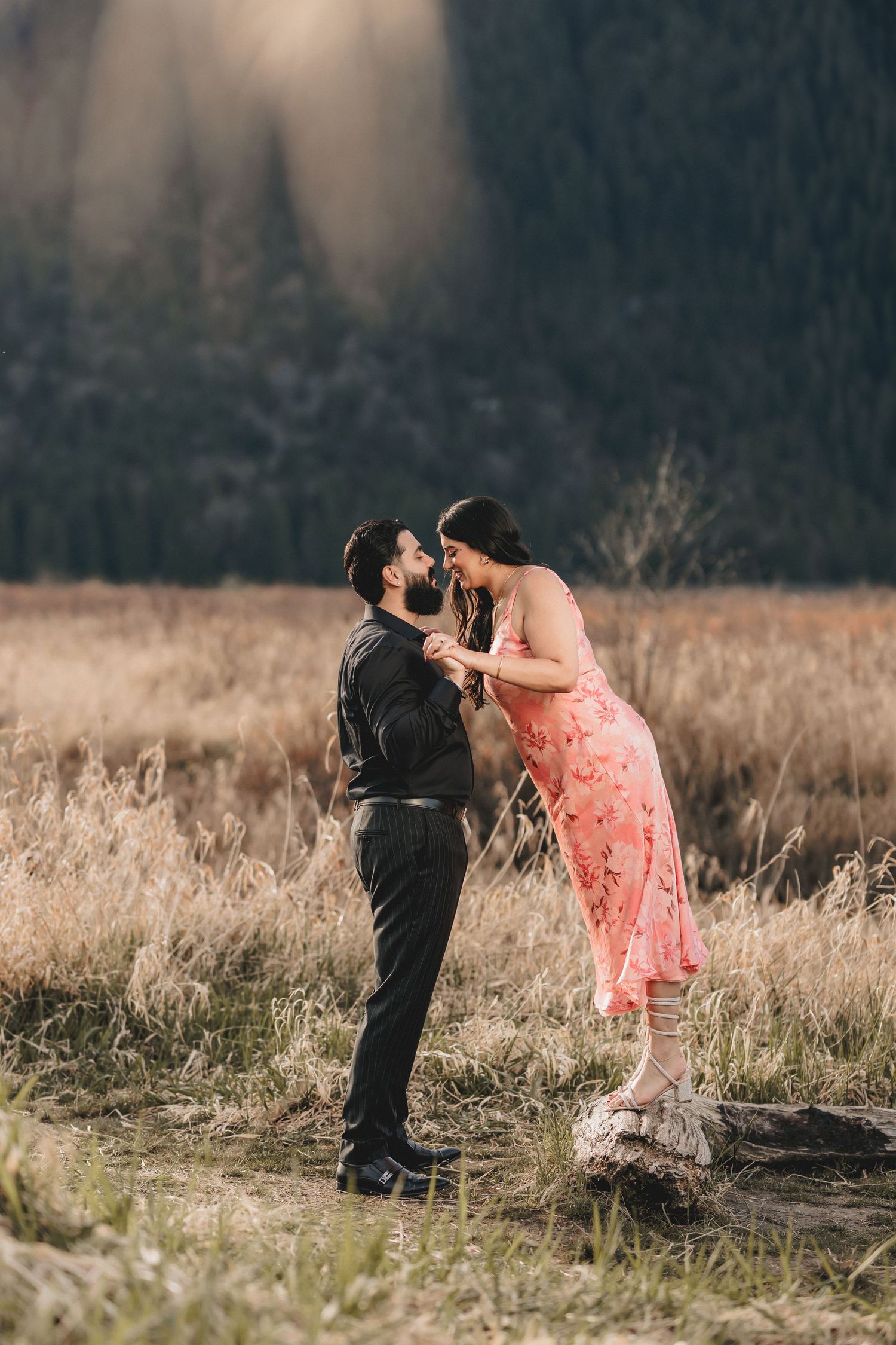 A couple in contrasting outfits dances playfully in a golden field with mountains behind them.