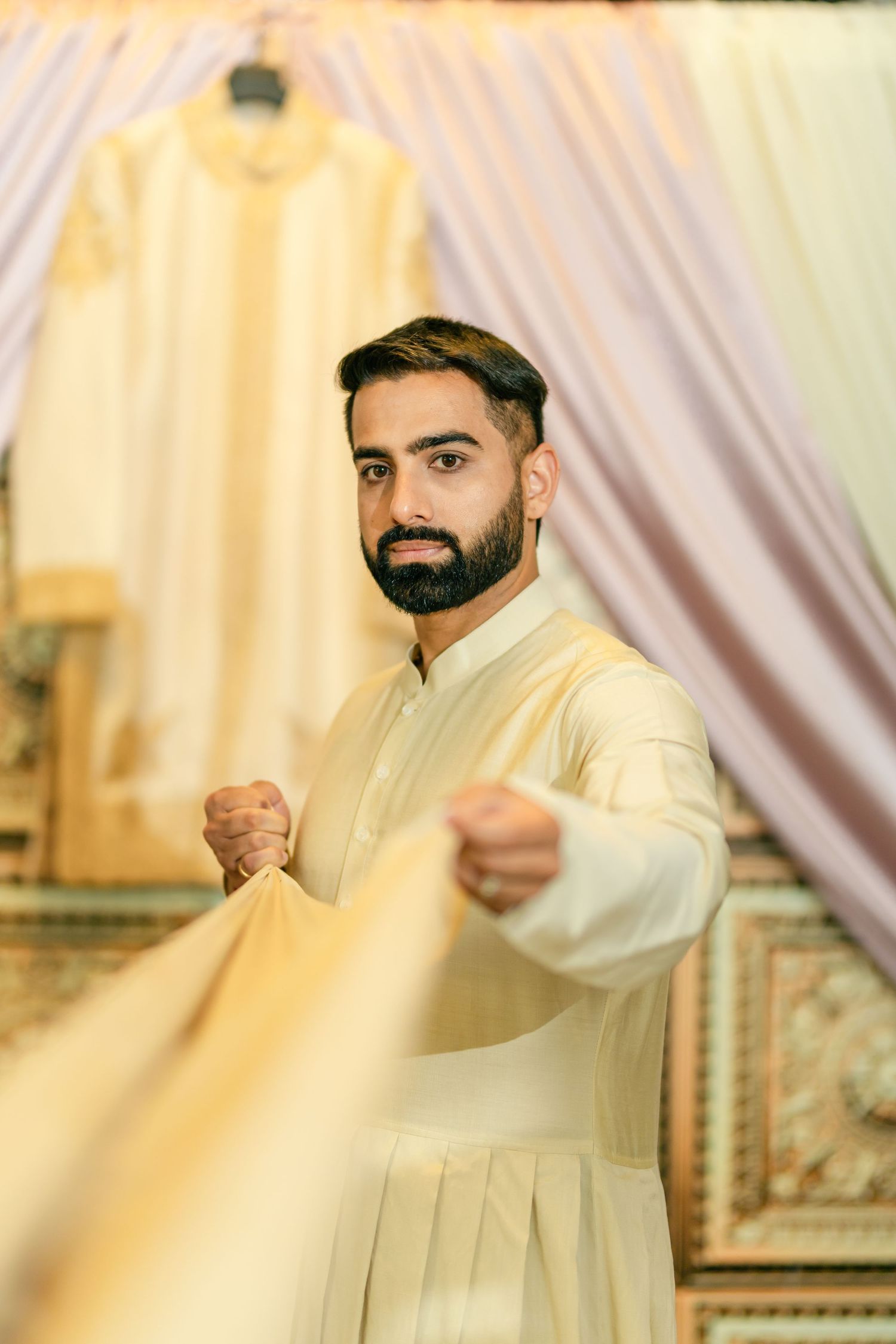 Man in cream colored traditional outfit striking playful poses against light backdrop.