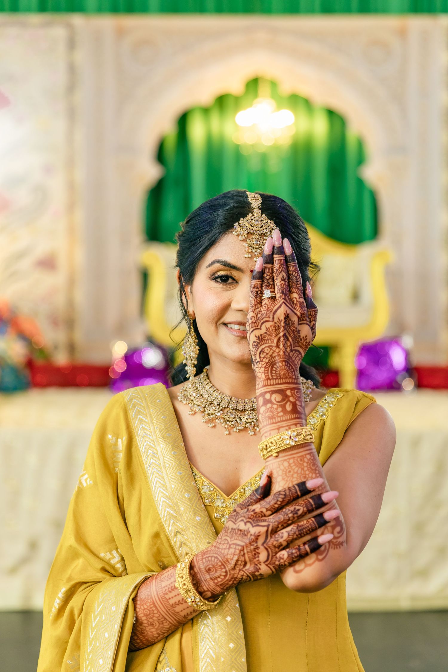 Person in yellow dress shows intricate henna designs on hands while wearing traditional Indian jewelry.
