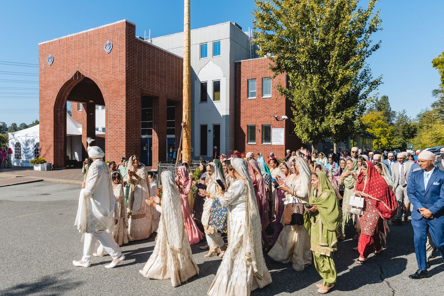 Wedding procession walking along a brick building exterior on a sunny autumn day.
