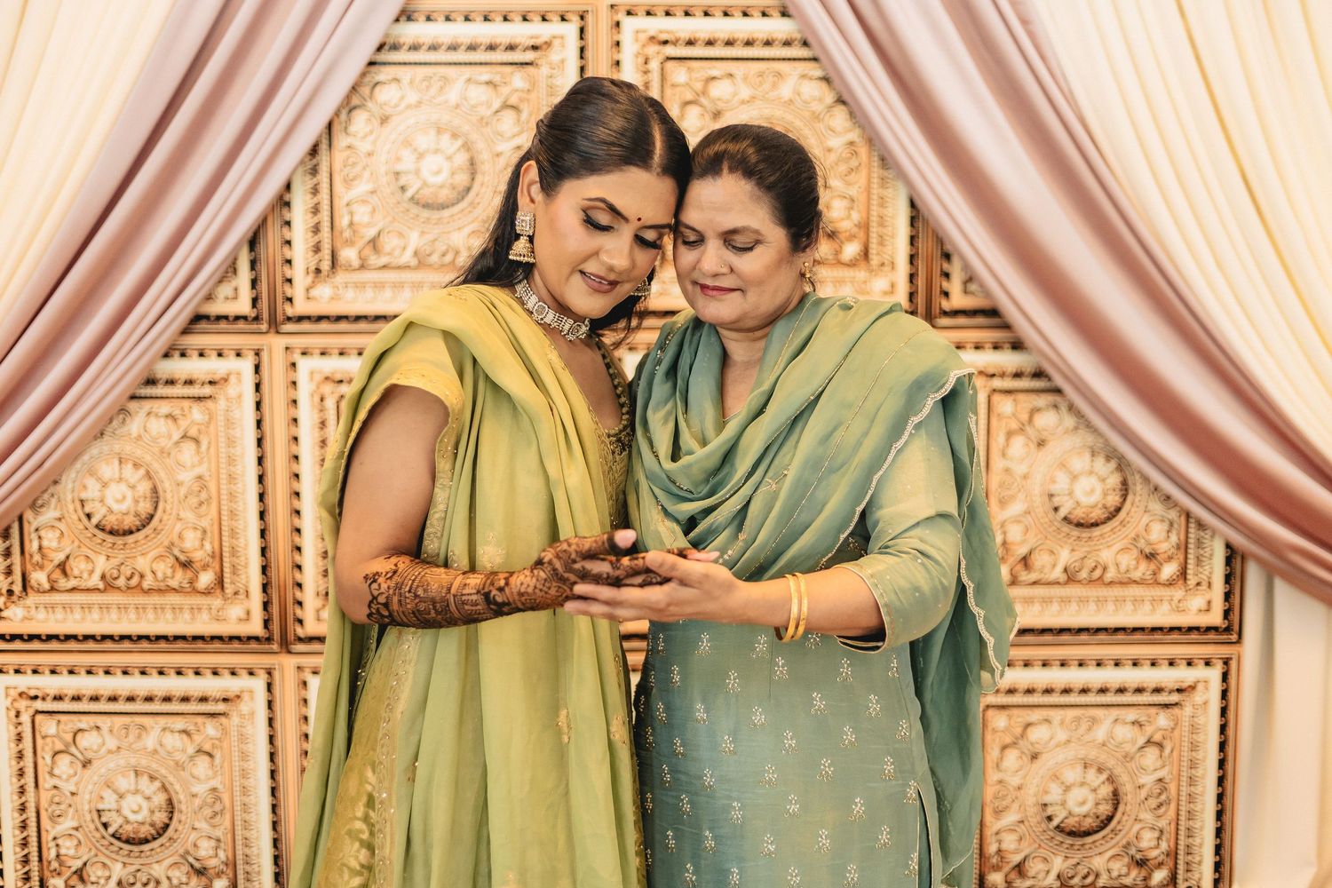 Two women in traditional saris look at a mobile phone together against an ornate gold-patterned backdrop.