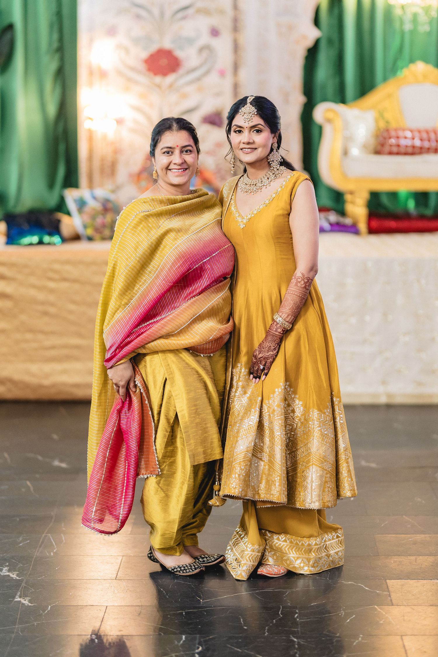 Two people wearing traditional Indian outfits in yellow and red pose together on a dark floor.