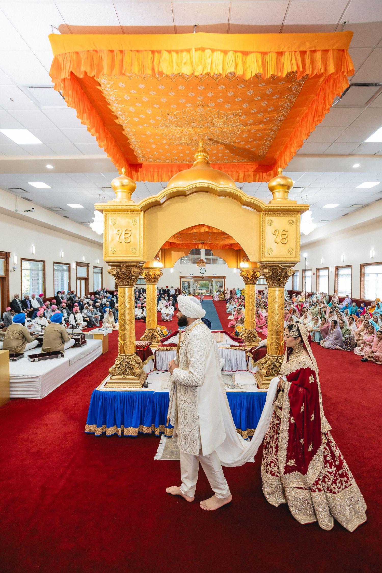 Traditional Sikh wedding ceremony taking place in a gurdwara with ornate orange and gold decorations and red carpeting.