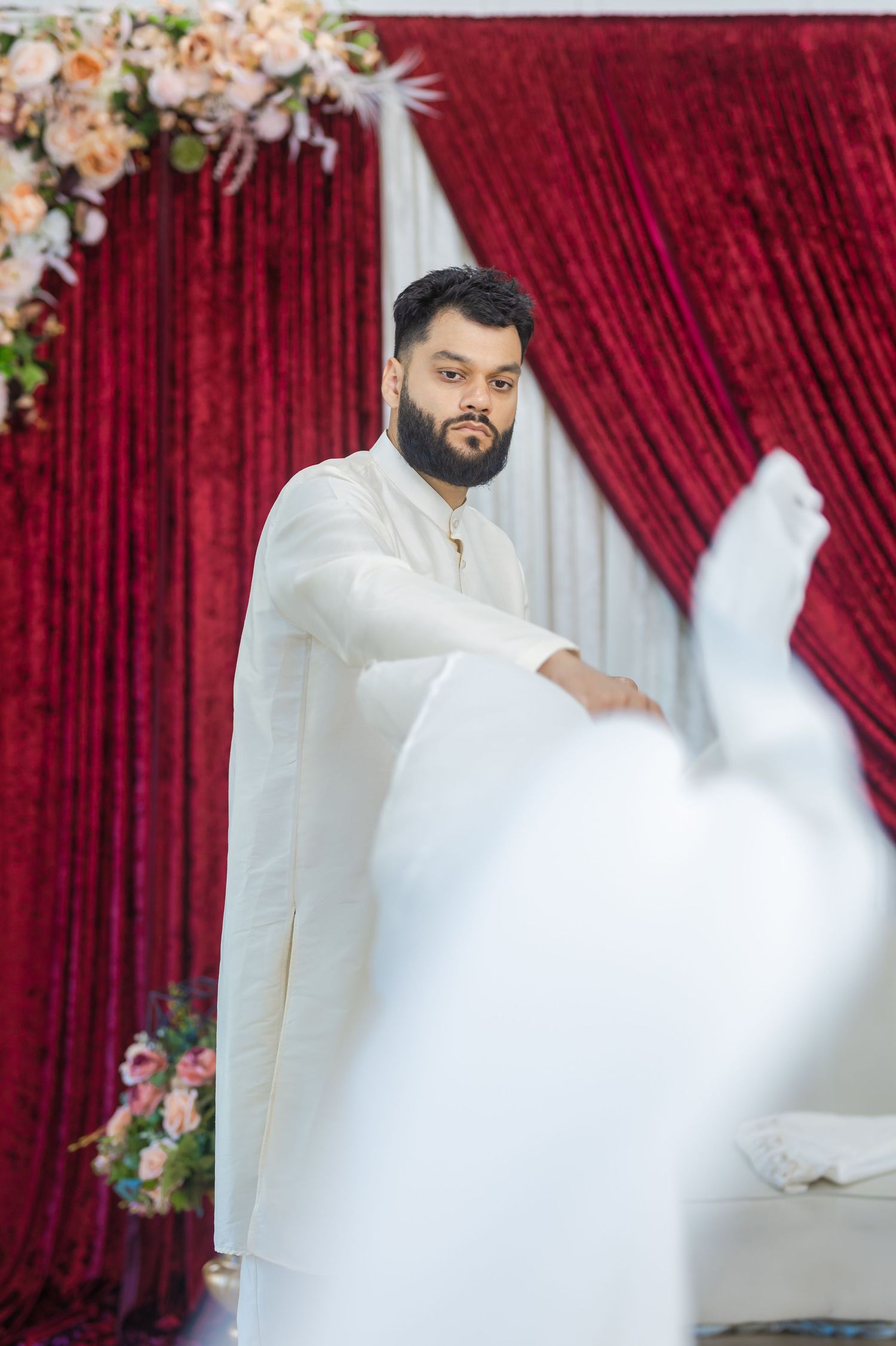 Person in white traditional attire poses against a red velvet backdrop with floral decorations.